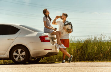 Young couple traveling on the car in sunny summer's day. Woman and man drinking coffee sitting on the trunk, stopping on the way to the sea. Concept of relationship, vacation, summer, holiday, weekend.